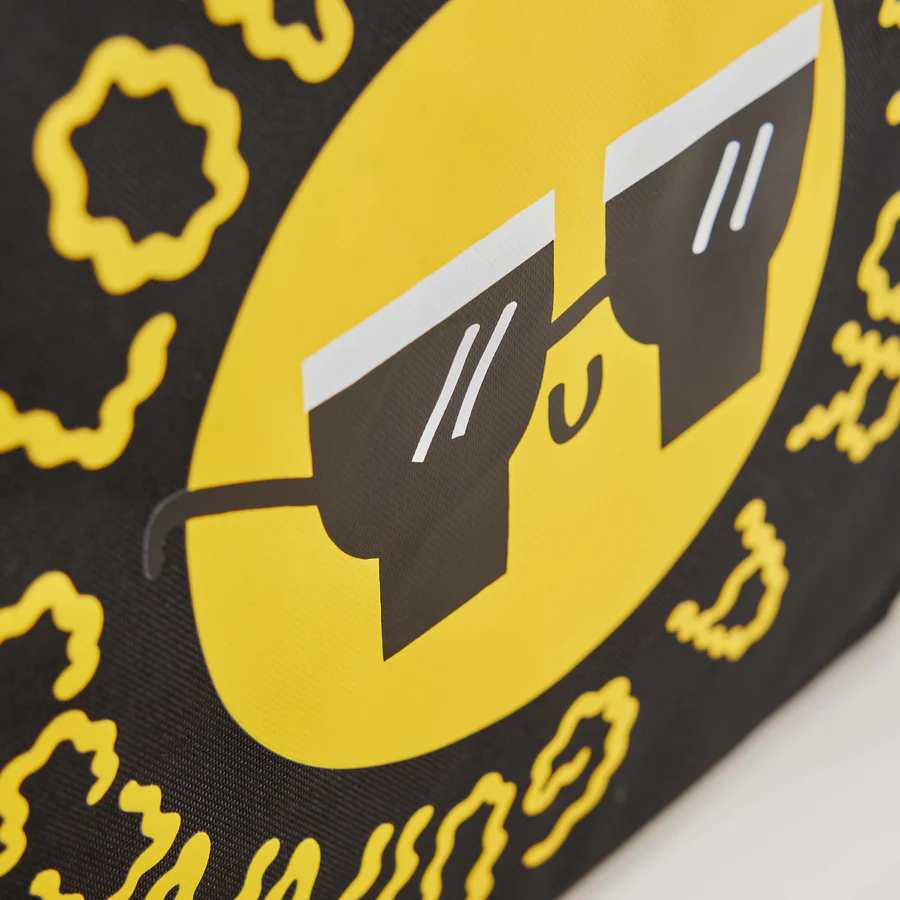 Close-up of a yellow smiley face with black sunglasses, inspired by Fatti Burke, on black fabric with yellow squiggles—featured on the Guinness x Fatti Burke Sun Cooler Bag from Guinness Webstore UK.