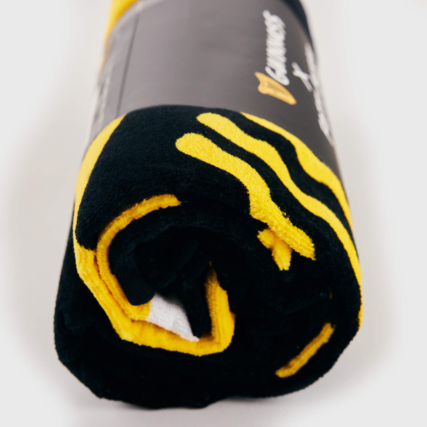 The Guinness x Fatti Burke Sun Beach Towel, a black and yellow cotton towel featuring a branded label, is rolled up and displayed on a white surface.