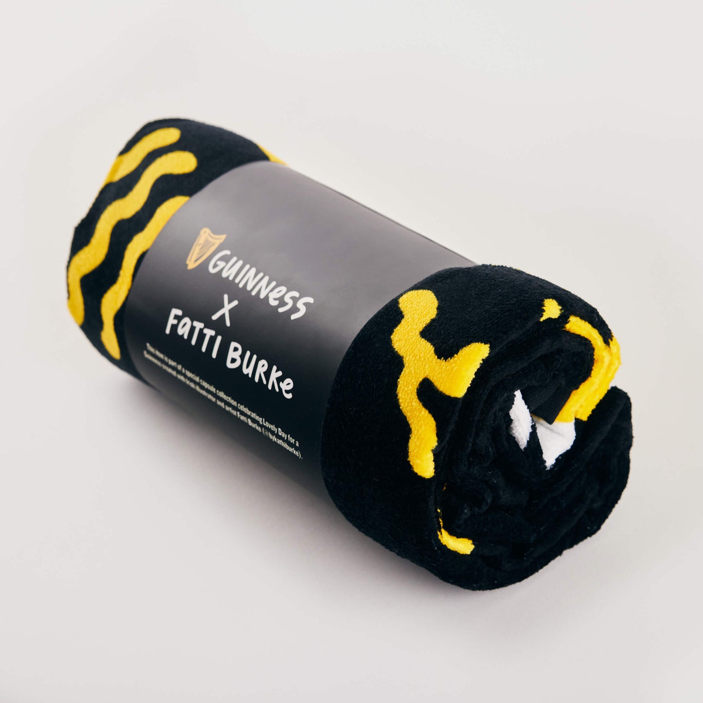The Guinness x Fatti Burke Sun Beach Towel, in black and yellow with a branded label, is rolled up and displayed on a light background.