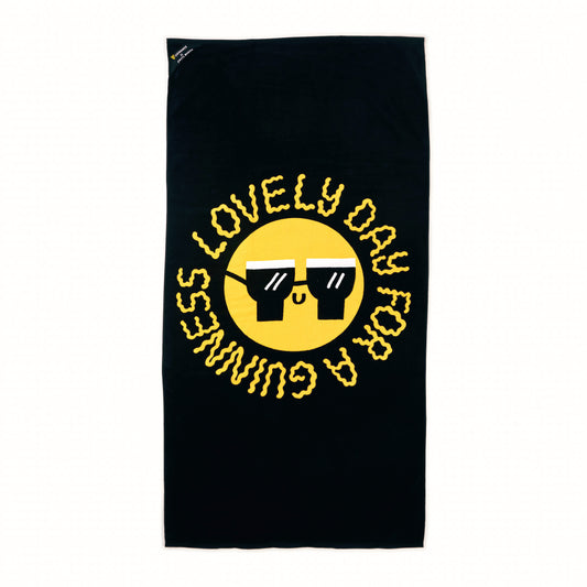 The Guinness x Fatti Burke Sun Beach Towel by Guinness features a large yellow smiling face with sunglasses and the phrase "LOVELY DAY FOR A GUINNESS" in bold yellow letters on a black background.