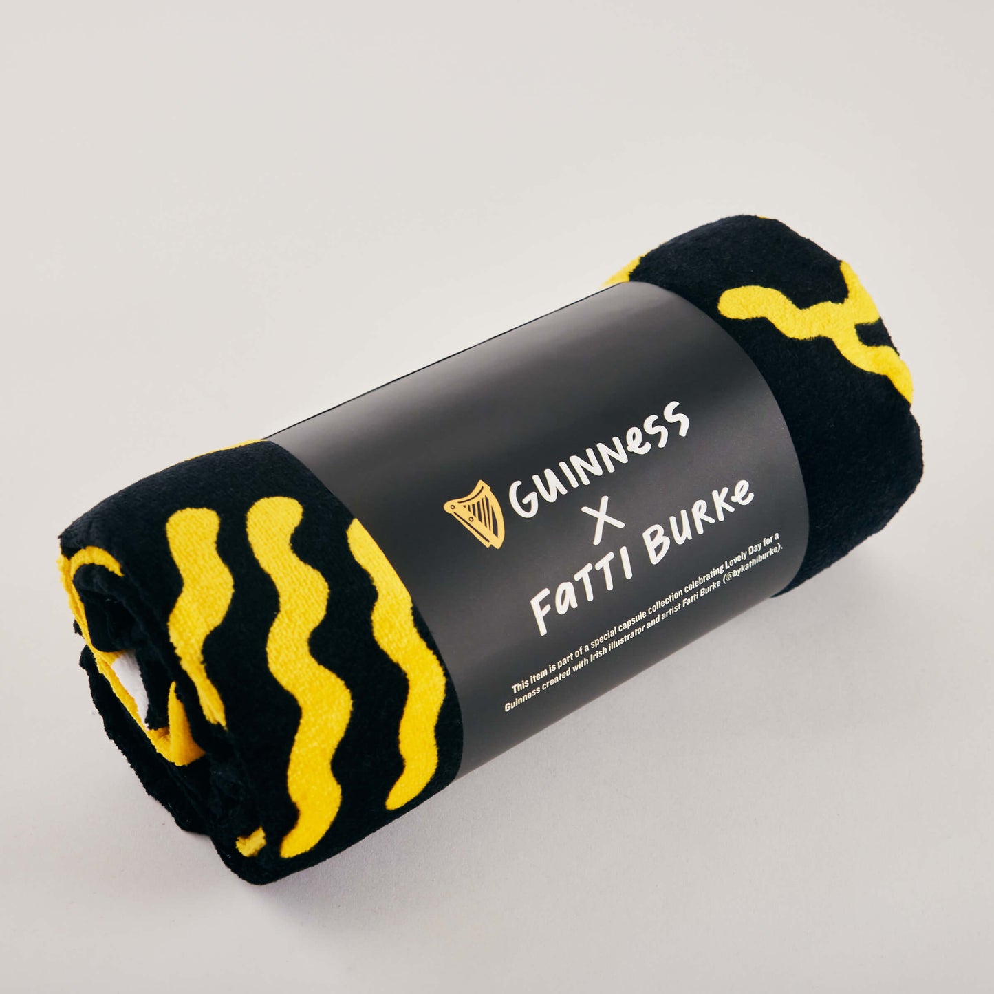 The Guinness x Fatti Burke Sun Beach Towel, featuring a black and yellow pattern, is rolled up with its label and displayed on a plain light surface.