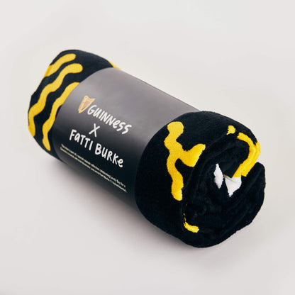 The Guinness x Fatti Burke Sun Beach Towel, in black and yellow with a branded label, is rolled up and displayed on a light background.