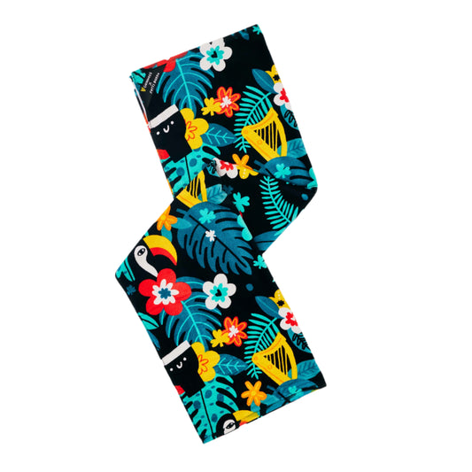 The Guinness x Fatti Burke Hawaiian Beach Towel by Guinness features a tropical leaf and flower pattern with toucans—including the Guinness Toucan—and hidden black cat faces on a dark background.